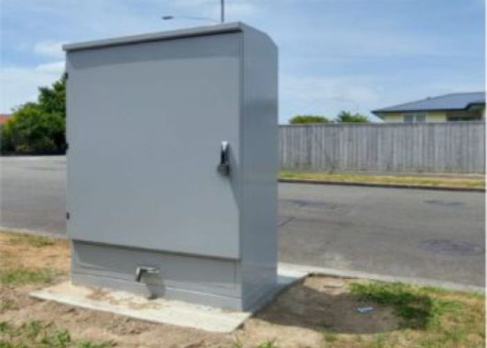 Grey kiosk with pump station inside on grass berm beside road blue sky